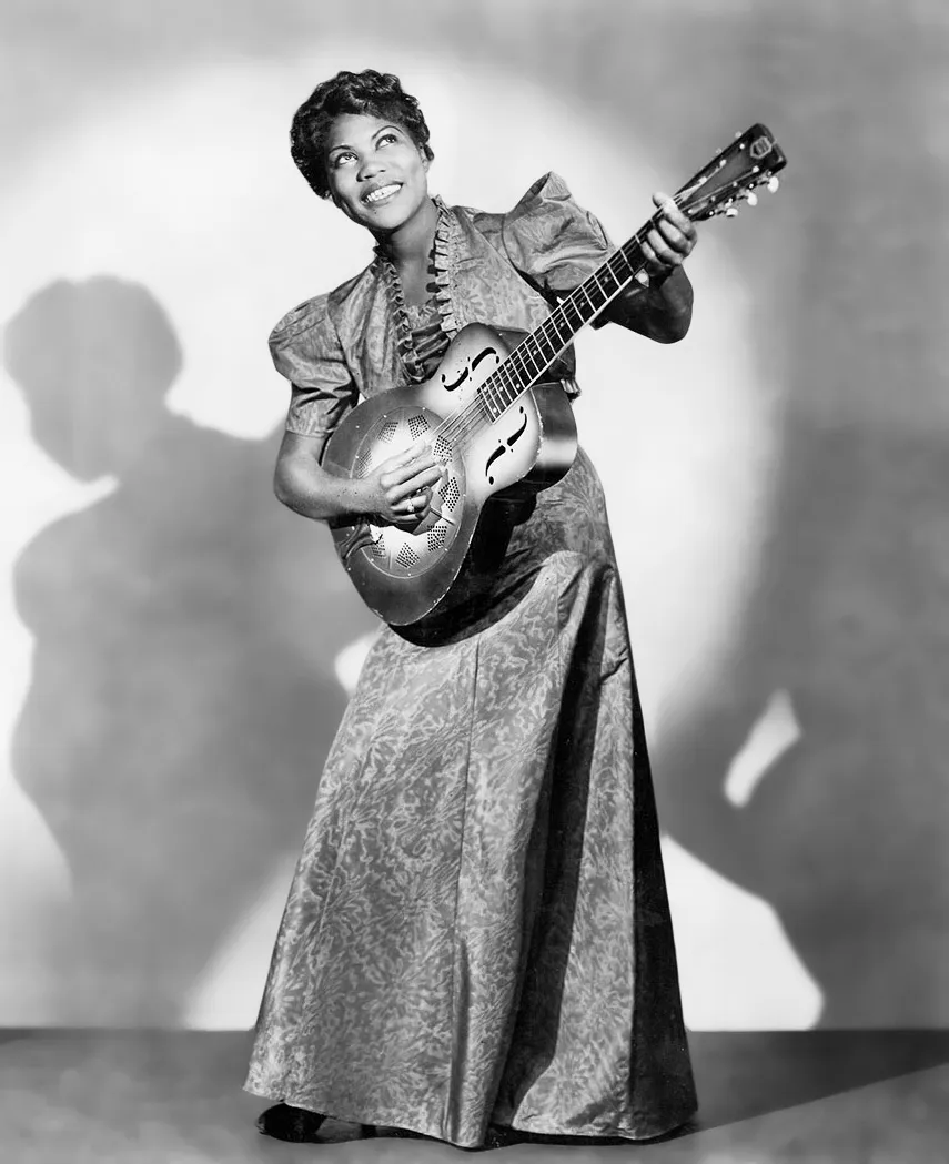 Full-length black-and-white publicity photograph of Sister Rosetta Tharpe standing beside a resonator guitar in 1938.
