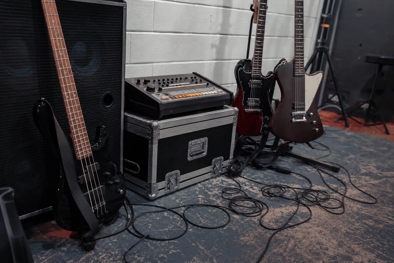 Conceptual editorial photograph of a bass, two electric guitars, a generic early drum machine, amplifier, road case, and cables in an empty rehearsal room.