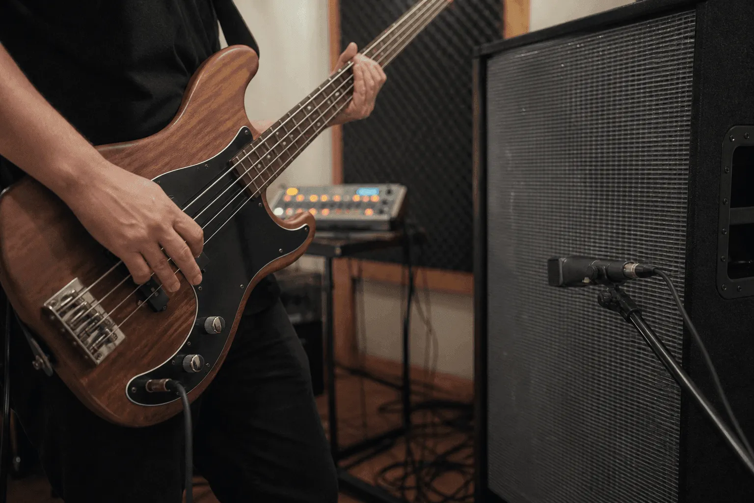Conceptual editorial photograph showing an anonymous musician's hands playing bass beside a miked speaker cabinet, with a generic drum machine behind.
