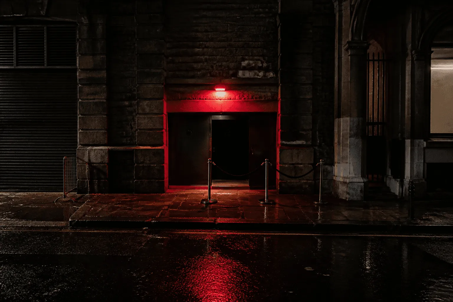 Conceptual editorial photograph of an anonymous narrow club entrance under a red light, with barriers and reflections on rain-wet pavement.