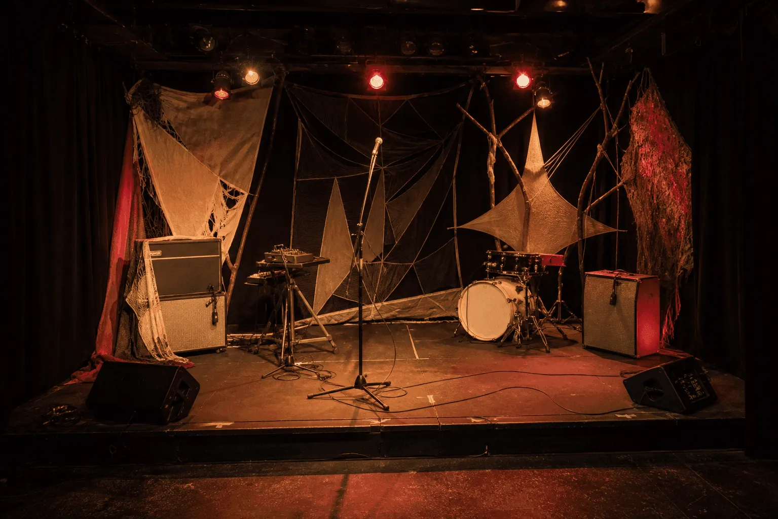 Conceptual editorial photograph of a tiny empty club stage with microphone, amplifiers, a compact machine stand, cables, black curtains, and handmade fabric decoration.